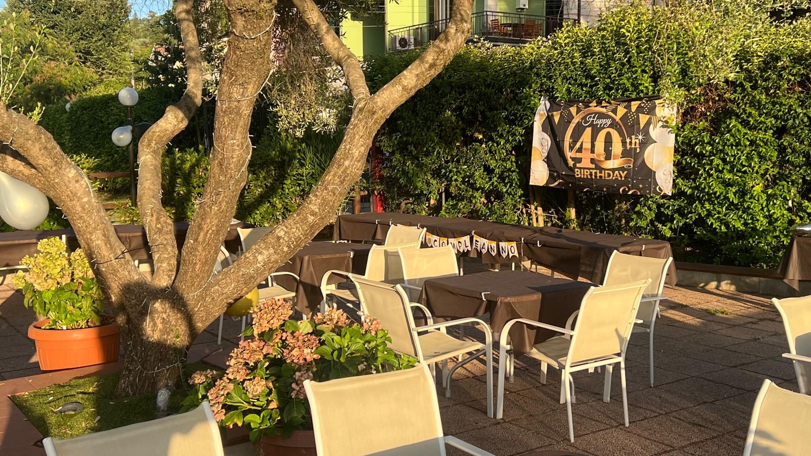 Dehors con tavoli dalle tovaglie marroni, sedie bianche e striscione Happy 40th Birthday sotto un ulivo con lucine